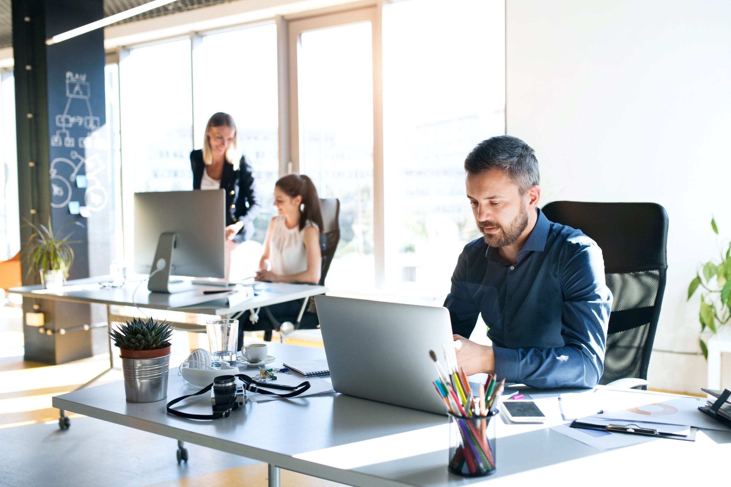 Mann arbeitet am Laptop und zwei Frauen sind im Hintergrund zu sehen